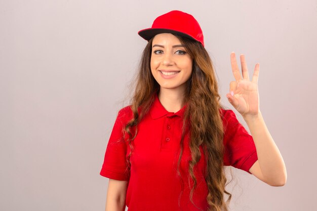 Junge Lieferfrau, die rotes Poloshirt und Kappe lächelnd freundlich tut ok Zeichen über lokalisierten weißen Hintergrund trägt