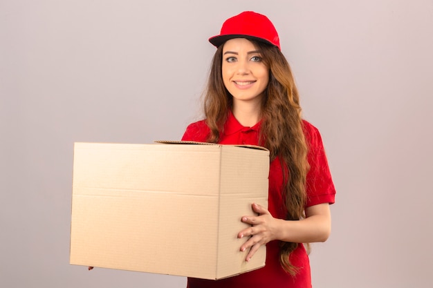 Junge Lieferfrau, die rotes Poloshirt und Kappe trägt, die mit Pappkarton steht, der fröhlich über lokalisiertem weißem Hintergrund lächelt