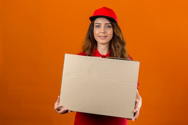 Junge Lieferfrau, die rotes Poloshirt und Kappe trägt, die mit Pappkarton steht, der zuversichtlich über lokalisiertem orange Hintergrund schaut