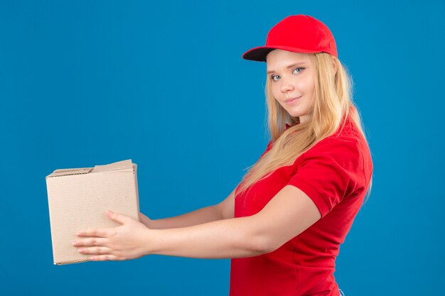 Junge Lieferfrau, die rotes Poloshirt und Kappe trägt, die Pappkarton zu einem Kunden geben, der zuversichtlich über lokalisierten blauen Hintergrund schaut