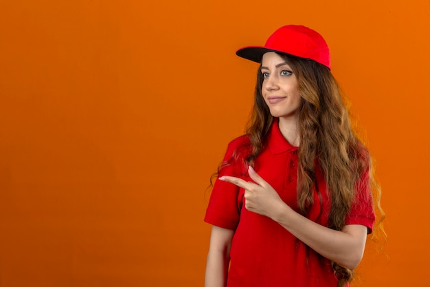 Junge Lieferfrau, die rotes Poloshirt und Kappe trägt, die zur Seite zeigen, um Kopienraum über lokalisiertem orangefarbenem Hintergrund zu präsentieren