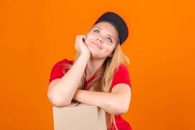 Kostenloses Foto junge lieferfrau, die rotes poloshirt und lächelnde kappe trägt, die wartende hand auf wange hält, während sie es mit einem anderen verträumten blick der gekreuzten hand über lokalisiertem orangefarbenem hintergrund unterstützt