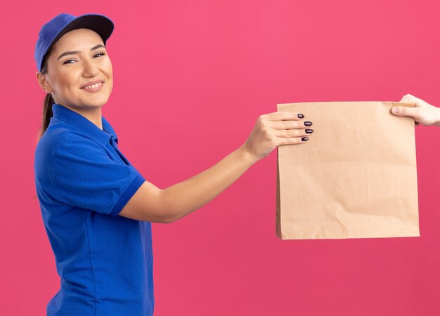 Junge Lieferfrau in blauer Uniform und Kappe, die Papierpaket zu einem Kunden lächelnd freundlich stehend über rosa Wand gibt