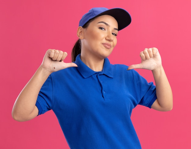 Junge Lieferfrau in blauer Uniform und Kappe glücklich und zuversichtlich, mit Daumen auf sich selbst stehend über rosa Wand zeigend