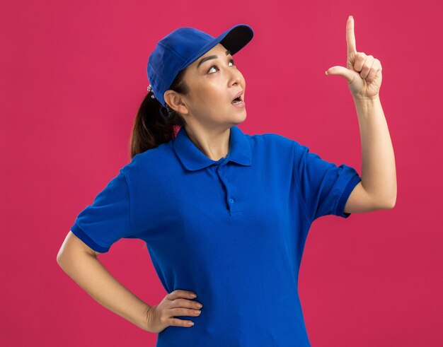 Junge Lieferfrau in blauer Uniform und Mütze, die mit dem Zeigefinger nach oben zeigt und neugierig nachdenkt