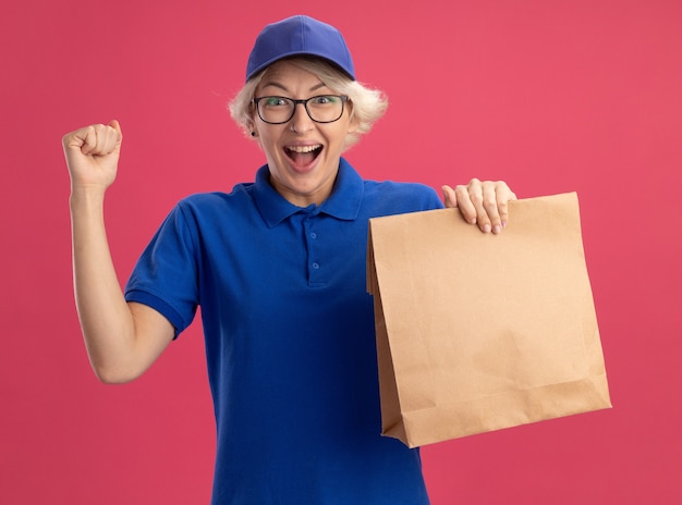 Junge Lieferfrau in der blauen Uniform und in der Kappe, die die Brille hält, die Papierpaket hält, das Faust glücklich und aufgeregt über rosa Wand erhebt