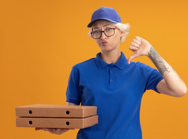 Junge Lieferfrau in der blauen Uniform und in der Kappe, die die Brille hält, die Pizzaschachteln hält, missfiel, Daumen über orange Wand unten zeigend