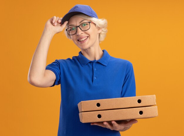 Junge Lieferfrau in der blauen Uniform und in der Kappe, die Gläser hält, die Pizzaschachteln halten, die zuversichtlich lächeln, ihre Kappe über orange Wand fixierend