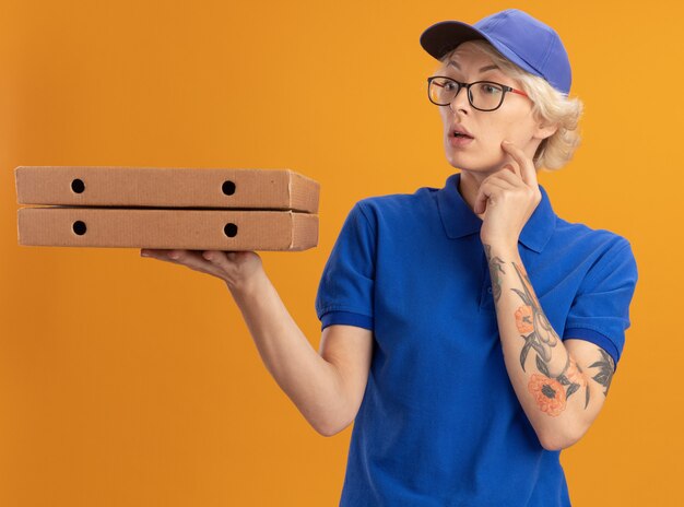 Junge Lieferfrau in der blauen Uniform und in der Kappe, die Gläser tragen, die Pizzakästen halten, die beiseite über orange Wand schauen