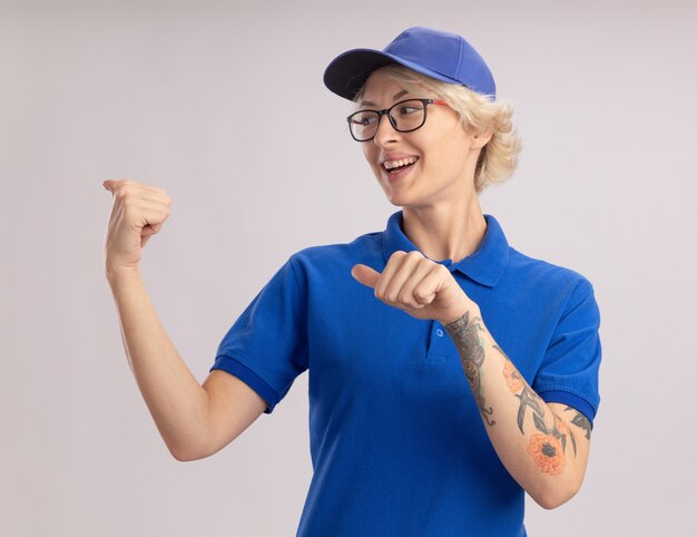 Junge Lieferfrau in der blauen Uniform und in der Kappe, die mit den Fingern zur Seite zeigt, die fröhlich über weißer Wand stehend lächelt