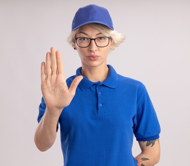 Junge Lieferfrau in der blauen Uniform und in der Kappe, die mit ernstem Gesicht schauen, das offene Hand zeigt, die Stoppgeste macht, die über weißer Wand steht