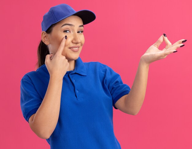 Junge Lieferfrau in der blauen Uniform und in der Kappe, die mit Zeigefinger auf ihr Auge zeigt, macht Geldgeste mit den Fingern, die über rosa Wand stehen