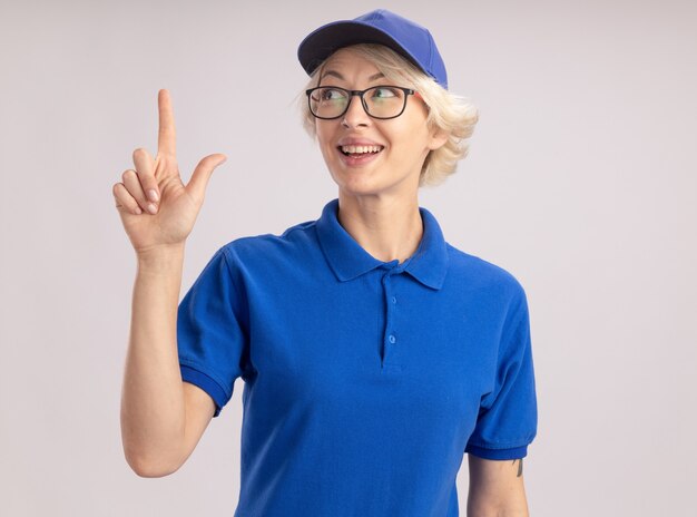 Junge Lieferfrau in der blauen Uniform und in der Kappe, die oben lächelnd zuversichtlich zeigt, mit Zeigefinger oben stehend über weißer Wand