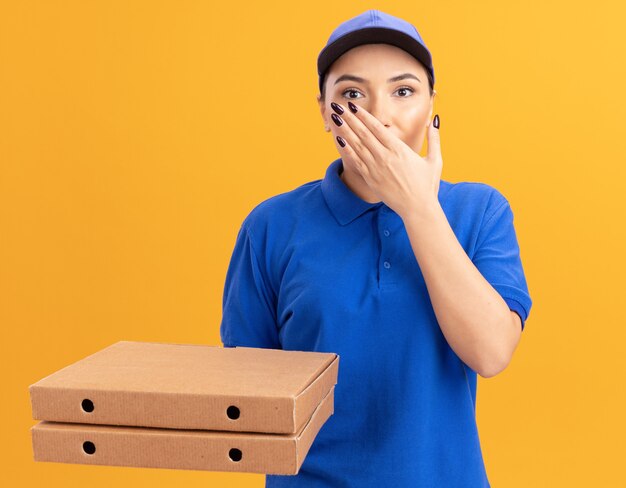 Junge Lieferfrau in der blauen Uniform und in der Kappe, die Pizzaschachteln betrachten, die vorne betrachtet werden, schockiert, Mund mit Hand, die über orange Wand steht