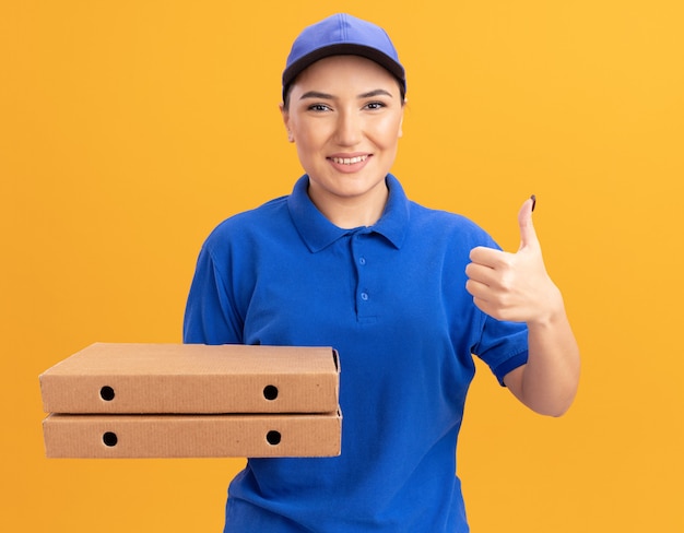 Junge Lieferfrau in der blauen Uniform und in der Kappe, die Pizzaschachteln hält, die vorne lächelnd zuversichtlich zeigen, Daumen hoch stehend über orange Wand