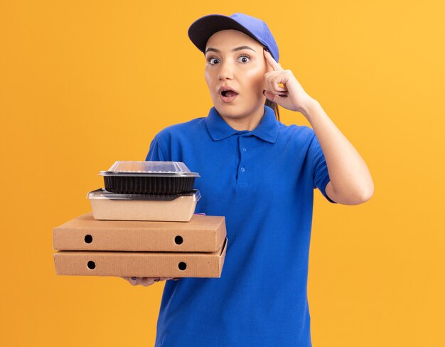 Junge Lieferfrau in der blauen Uniform und in der Kappe, die Pizzaschachteln und Lebensmittelverpackungen hält, die nach vorne zeigen mit Zeigefinger auf ihre Schläfe schauen verwirrt über orange Wand stehend