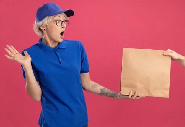 Junge Lieferfrau in der blauen Uniform und in der Kappe, die sich aufgeregt fühlen, während sie Papierpaket erhalten, das über rosa Wand steht