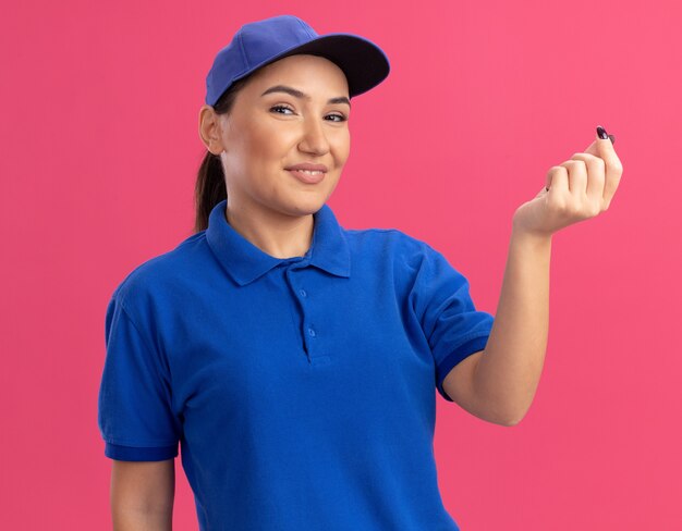 Junge Lieferfrau in der blauen Uniform und in der Kappe, die vorne schaut, macht Geldgeste, die Finger reibt, die sicher lächelnd über rosa Wand stehend lächeln