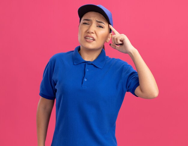 Junge Lieferfrau in der blauen Uniform und in der Mütze, die vorne betrachtet, verwechselt mit Finger nahe ihrer Schläfe, die über rosa Wand steht