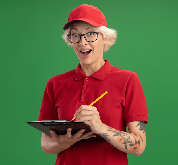 Junge Lieferfrau in der roten Uniform und in der Kappe, die Brille mit Zwischenablage mit leeren Seiten und Bleistift trägt, schreibt etwas, das glücklich und überrascht über grüner Wand steht
