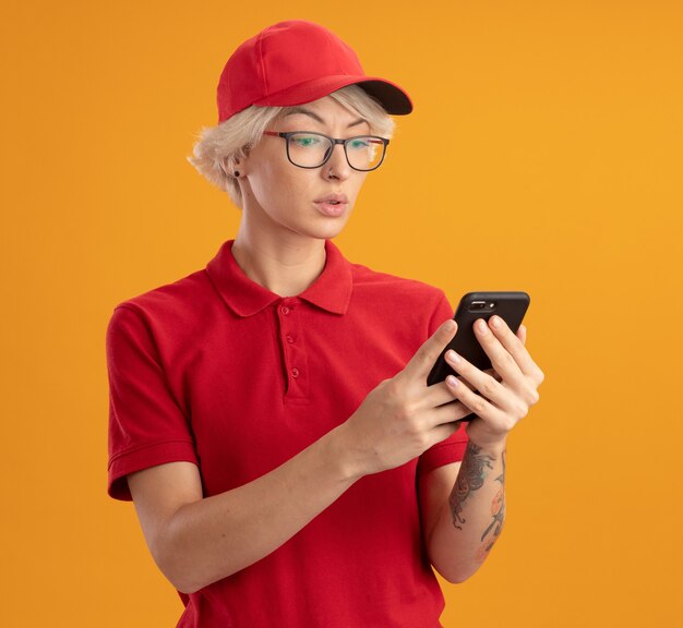 Junge Lieferfrau in der roten Uniform und in der Kappe, die Brille tragen, die ihr Smartphone mit ernstem Gesicht über orange Wand steht