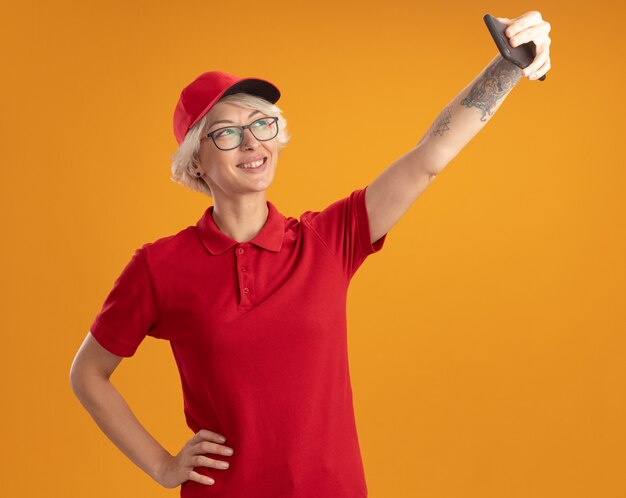 Junge Lieferfrau in der roten Uniform und in der Kappe, die Brille unter Verwendung des Smartphones tut, das freudig lächelndes Selfie über orange Wand steht