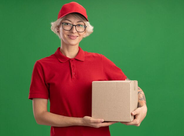 Junge Lieferfrau in der roten Uniform und in der Kappe, die die Brille hält, die Karton hält und lächelnd zuversichtlich über grüner Wand steht