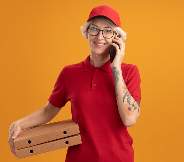 Junge Lieferfrau in der roten Uniform und in der Kappe, die die Brille hält, die Pizzakästen hält, die auf Handy sprechen, das fröhlich über orange Wand steht