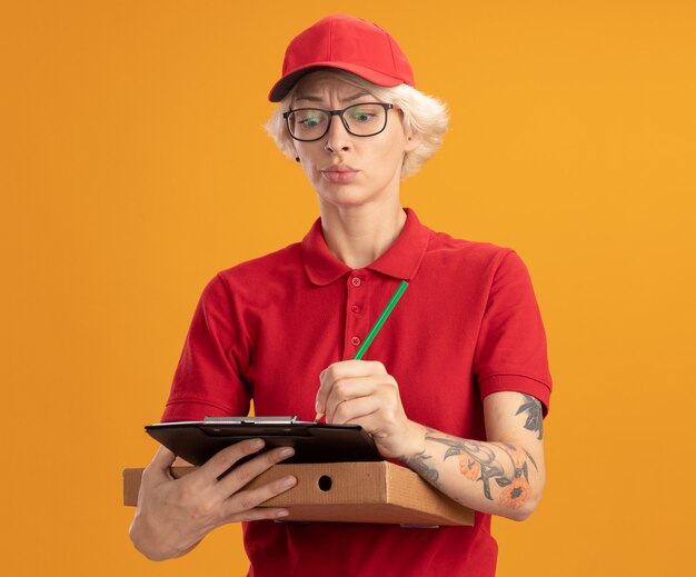 Junge Lieferfrau in der roten Uniform und in der Kappe, die die Brille hält, die Pizzaschachtel und Zwischenablage mit Bleistift hält, der etwas mit ernstem Gesicht schreibt, das über orange Wand steht