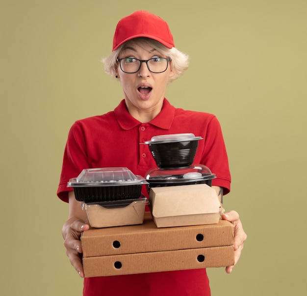 Junge Lieferfrau in der roten Uniform und in der Kappe, die Gläser hält, die Pizzaschachteln und Lebensmittelverpackungen halten, überrascht und erstaunt, über grüner Wand stehend