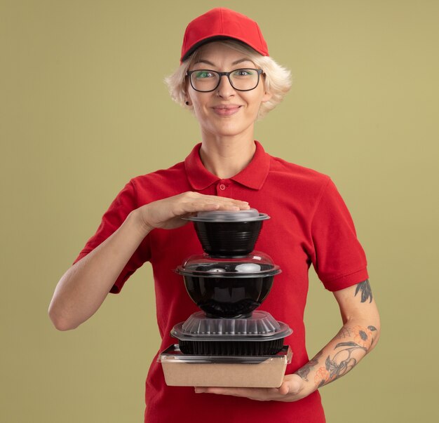 Junge Lieferfrau in der roten Uniform und in der Kappe, die Gläser tragen, die Stapel von Lebensmittelpaketen halten, die zuversichtlich über grüner Wand stehen