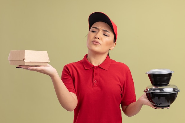 Junge Lieferfrau in der roten Uniform und in der Kappe, die Lebensmittelpakete halten, die verwirrt und unsicher stehen über grüner Wand stehen