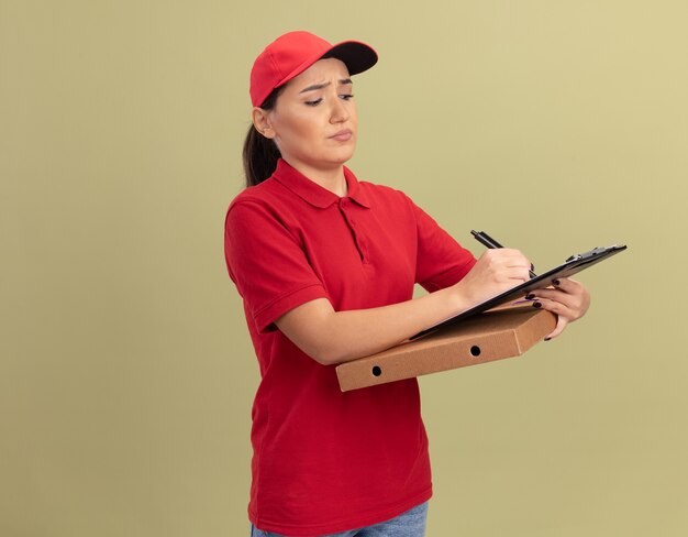 Junge Lieferfrau in der roten Uniform und in der Kappe, die Pizzaschachtel mit Klemmbrett und Bleistift hält, der mit ernstem Gesicht steht, das über grüner Wand steht