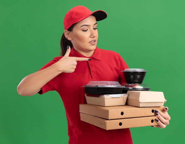 Junge lieferfrau in der roten uniform und in der kappe, die pizzaschachteln und lebensmittelverpackungen hält, die mit zeigefinger auf sie zeigen, die verwirrt sind, die über grüner wand stehen