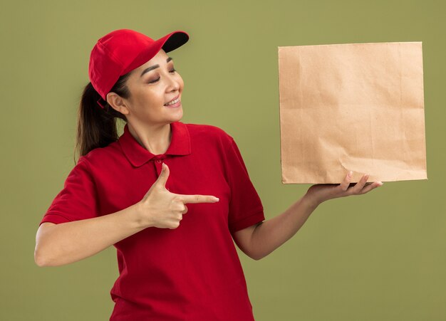 Junge Lieferfrau in roter Uniform und Mütze, die ein Papierpaket hält und mit dem Zeigefinger darauf zeigt, lächelt selbstbewusst über grüner Wand