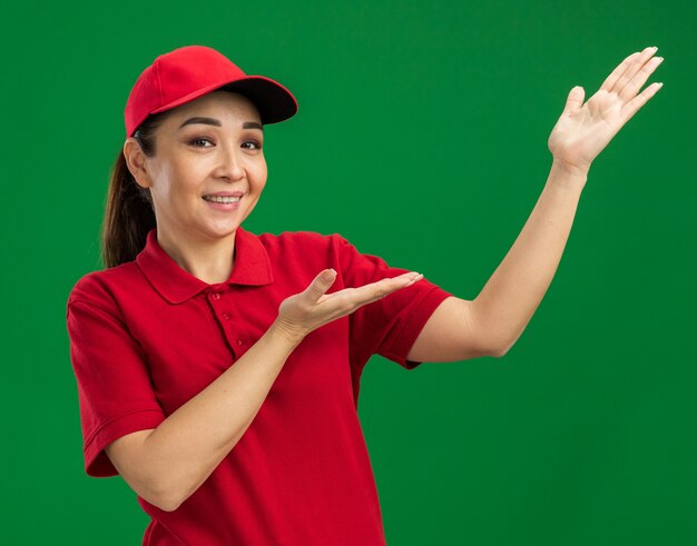 Junge Lieferfrau in roter Uniform und Mütze, die Kopienraum mit den Armen der Hände präsentiert, die glücklich und positiv lächeln