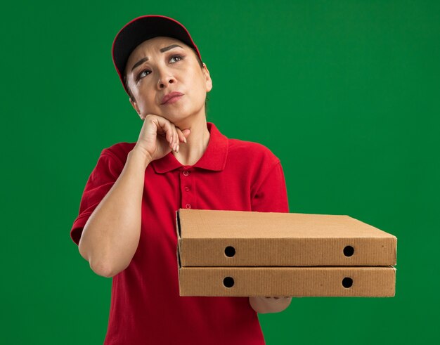 Junge Lieferfrau in roter Uniform und Mütze, die Pizzakartons hält und verwirrt aufschaut
