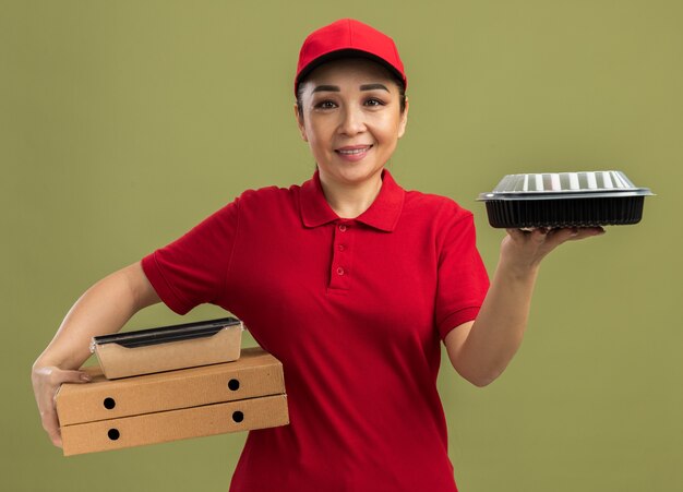 Junge Lieferfrau in roter Uniform und Mütze, die Pizzakartons und Lebensmittelpakete mit einem Lächeln im Gesicht hält, das über grüner Wand steht