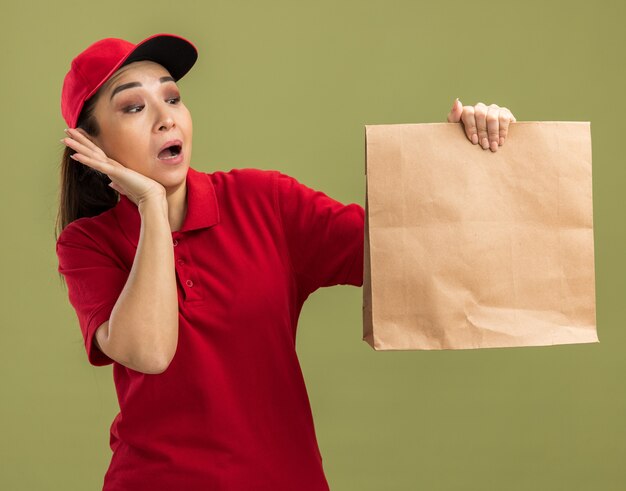 Junge Lieferfrau in roter Uniform und Mütze mit Papierpaket, die es erstaunt und überrascht betrachtet