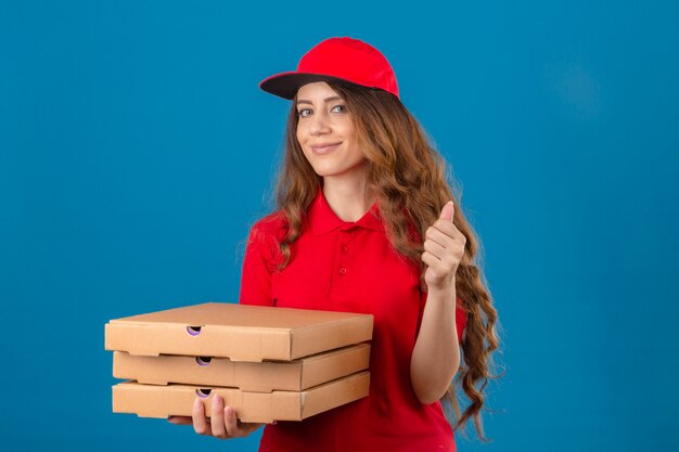 Junge Lieferfrau mit dem gelockten Haar, das rotes Poloshirt und Kappe mit dem Stapel lächelnder Pizzaschachteln trägt, zeigt Daumen oben über lokalisiertem blauem Hintergrund