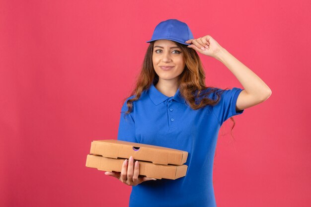 Junge Lieferfrau mit dem lockigen Haar, das blaues Poloshirt und Kappe trägt, die mit Pizzakästen salutiert, die zuversichtlich über lokalisiertem rosa Hintergrund suchen