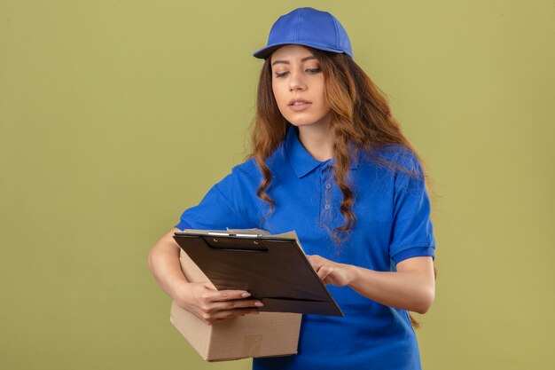 Junge Lieferfrau mit lockigem Haar, das blaues Poloshirt und Kappe trägt, die Klemmbrett in Händen mit ernstem Gesicht betrachtet, das auf Aufgabe über lokalisiertem grünem Hintergrund konzentriert ist