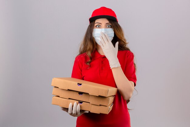 Junge Lieferfrau mit lockigem Haar, das rotes Poloshirt und Kappe in der medizinischen Schutzmaske und in den Handschuhen trägt, die mit Pizzakästen stehen, die überraschtes Abdecken des Mundes mit Hand über isoliertem weißem ba stehen