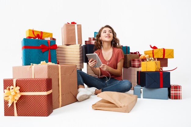 Junge lockige Frau, die auf Boden unter Geschenkboxen mit gerade geöffneter Geschenk-Tablette sitzt