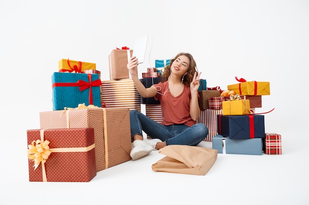 Junge lockige Frau, die auf Boden unter Geschenkboxen sitzt, die selfie mit Tablette machen