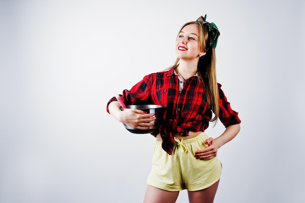 Kostenloses Foto junge lustige hausfrau in kariertem hemd und gelben shorts pin-up-stil mit kochtopf und küchenlöffel isoliert auf weißem hintergrund