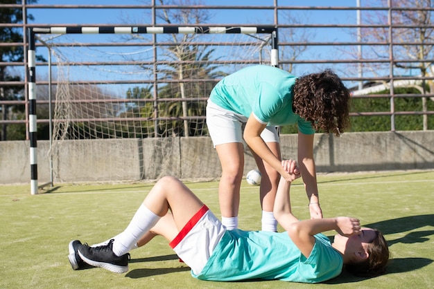 Junge Mädchen ruhen sich aus, nachdem sie im Fußballspiel gewonnen haben. Zwei sportliche Mädchen, die sich über den Sieg freuen, und ein Mädchen, das müde auf grünem Boden liegt, während ihre Freundin ihr hilft. Freundschaft, Mannschaftssportkonzept