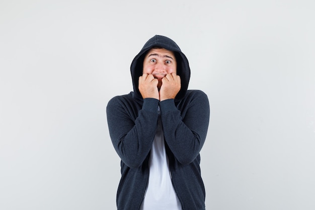 Junge männliche beißende Fäuste emotional in T-Shirt, Jacke und erschrocken aussehend. Vorderansicht.