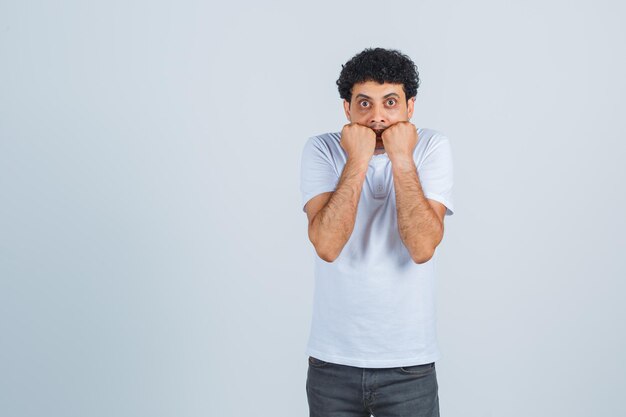 Junge männliche beißende Fäuste emotional in weißem T-Shirt, Hose und verängstigtem Blick. Vorderansicht.