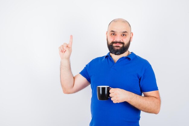 Junge männliche Holding Tasse beim Zeigen nach oben im blauen Hemd, Vorderansicht.